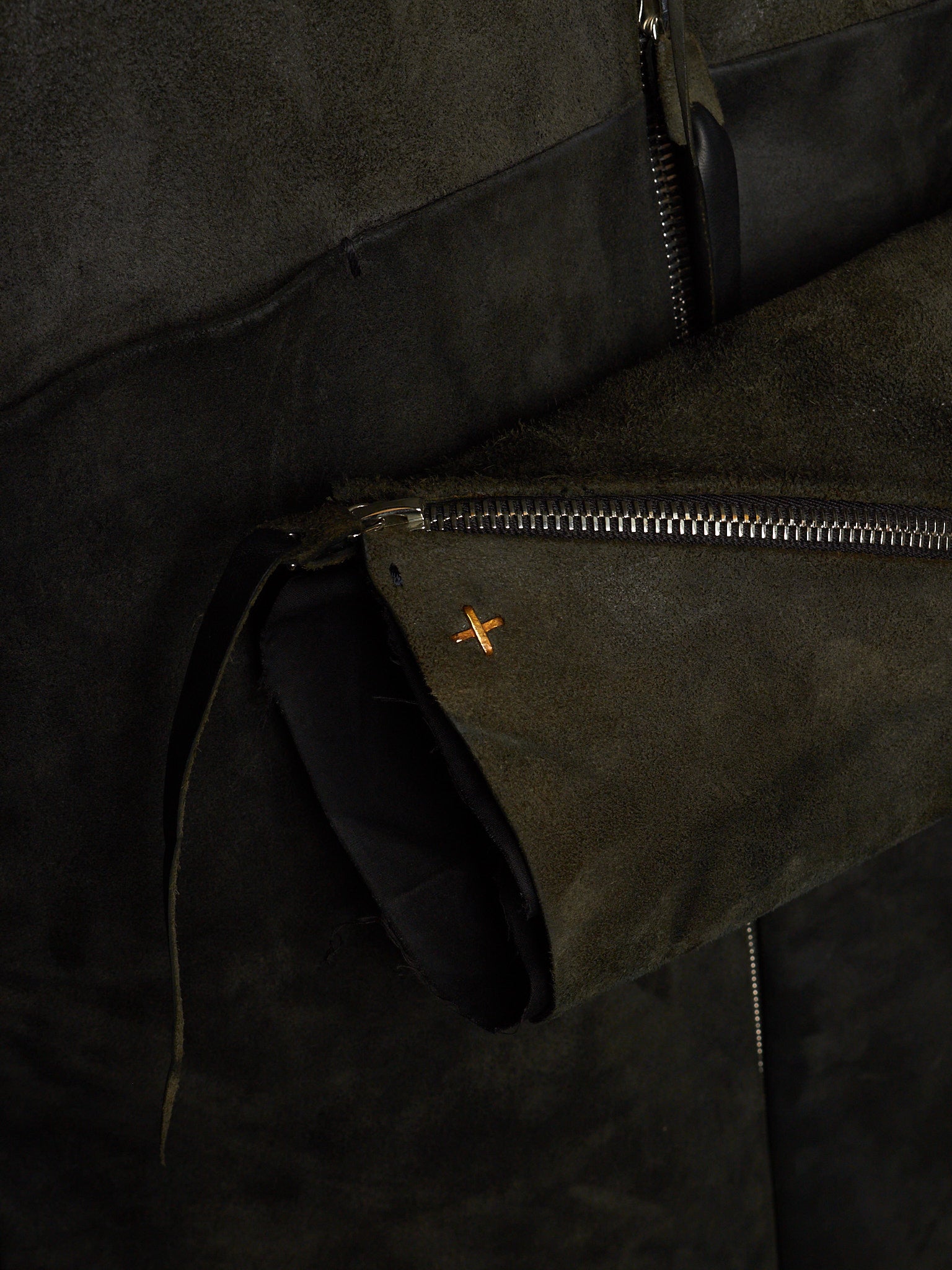 Close-up of a dark olive green suede jacket with a silver zipper and a small orange embroidered cross logo.