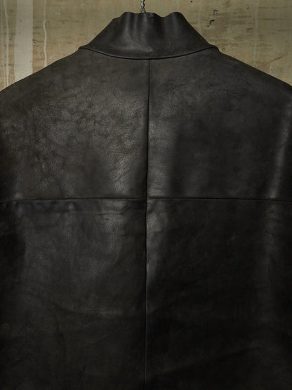 A close-up of a black leather jacket or coat hanging against a textured beige wall, showing the back panel with a high collar and a central vertical seam.