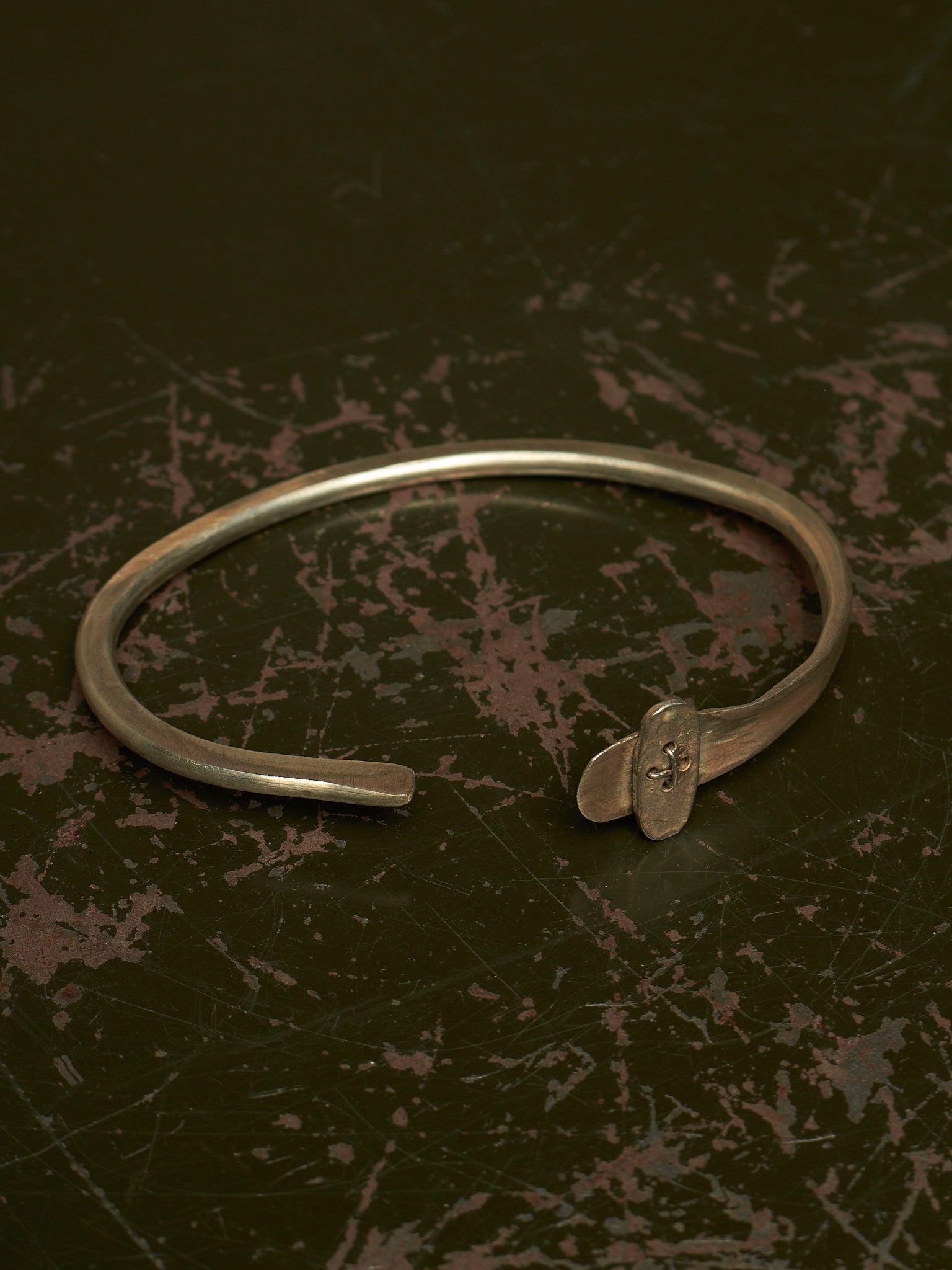 A silver-toned metal cuff bracelet with a small decorative element at the open end, photographed on a dark textured surface.