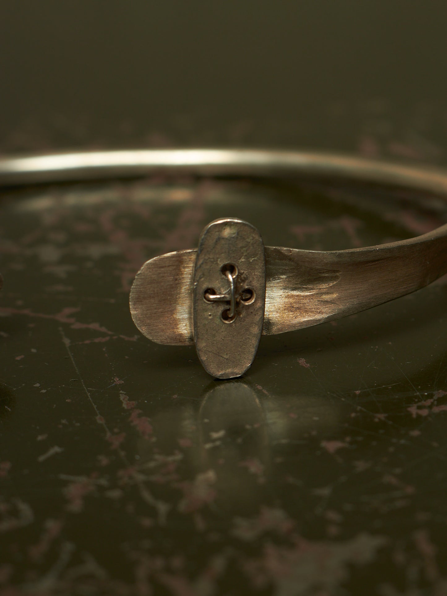 A close-up of a rustic silver-toned metal cuff bracelet featuring a central oval plate with a cutout fleur-de-lis design, resting on a dark reflective surface.