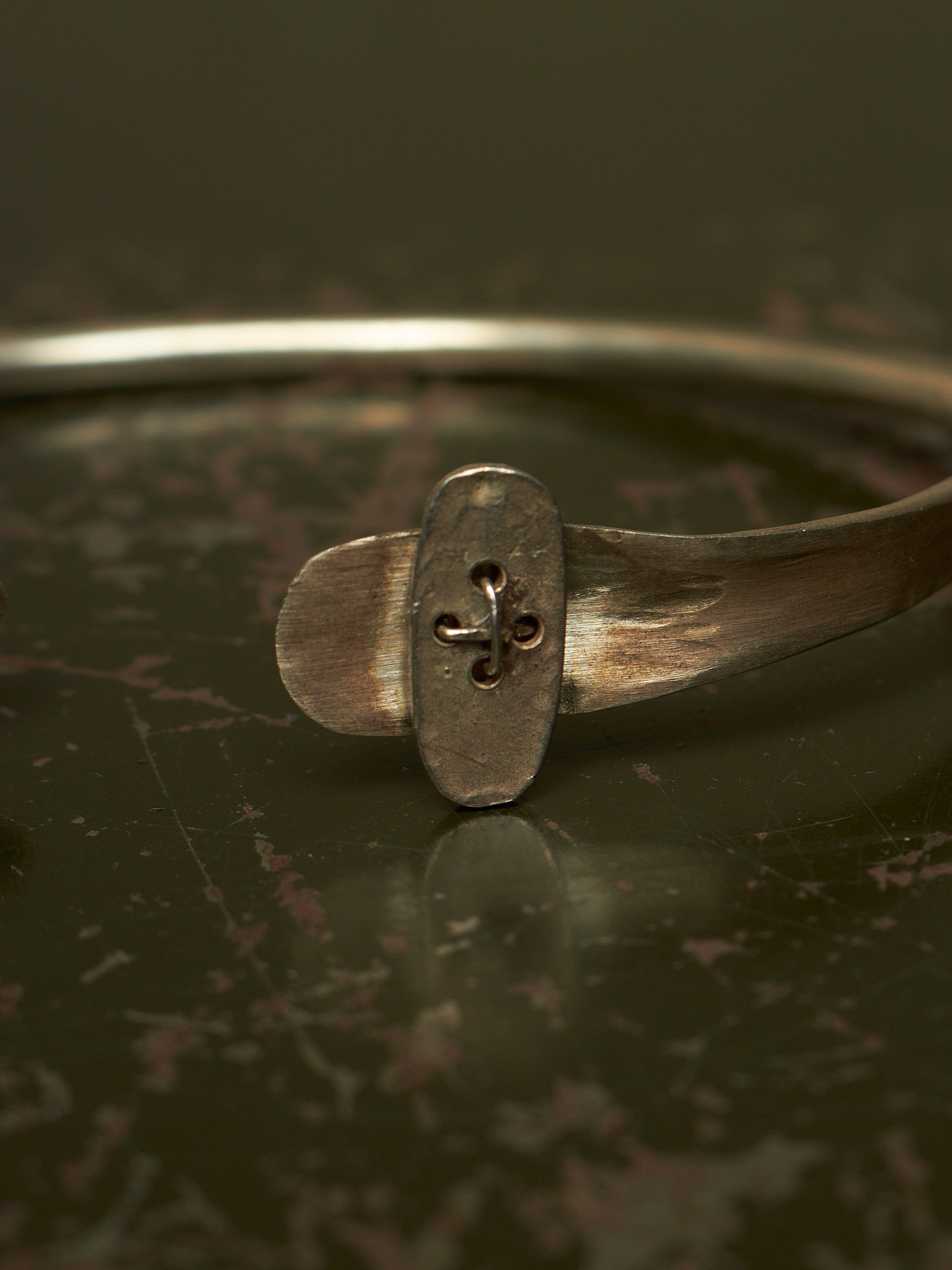 A close-up of a rustic silver-toned metal cuff bracelet featuring a central oval plate with a cutout fleur-de-lis design, resting on a dark reflective surface.