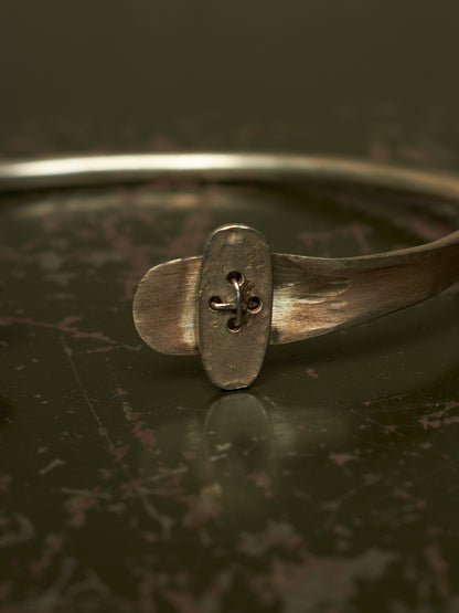 A close-up of a rustic silver-toned metal cuff bracelet featuring a central oval plate with a cutout fleur-de-lis design, resting on a dark reflective surface.