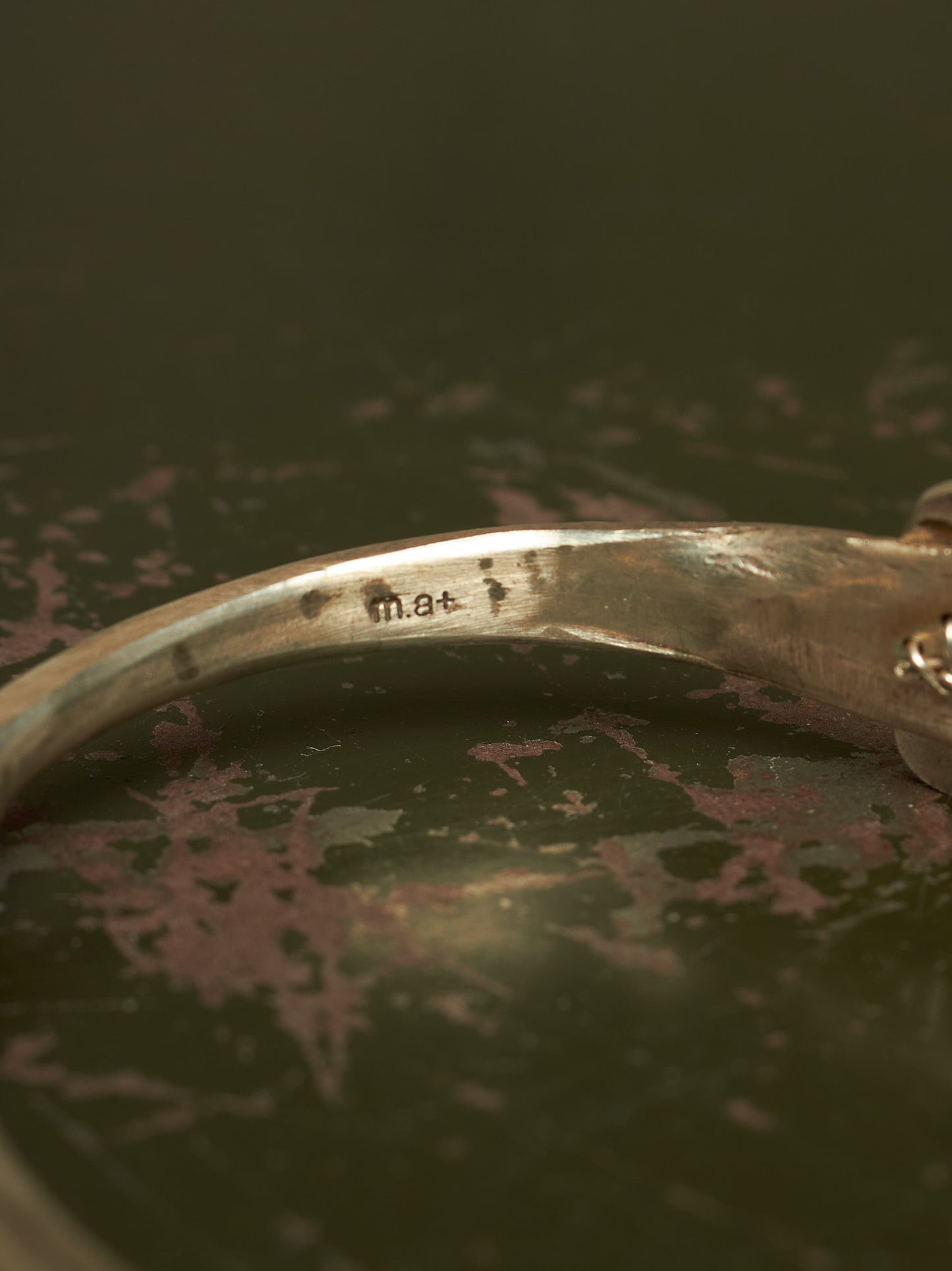 Close-up of a silver-toned metal bangle bracelet showing a stamped 'mat' mark inside the band, resting on a dark textured surface.
