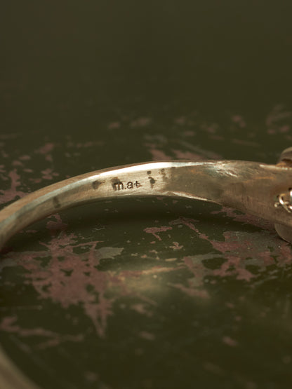 Close-up of a silver-toned metal bangle bracelet showing a stamped 'mat' mark inside the band, resting on a dark textured surface.