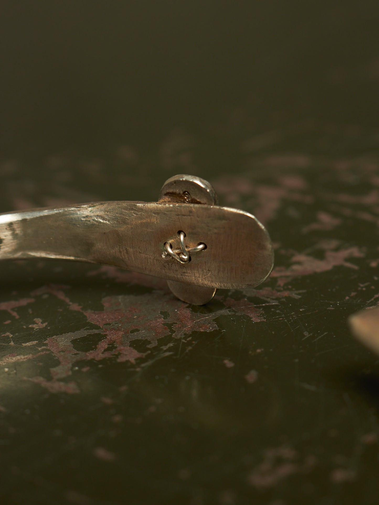 A close-up of a silver metal spoon handle with a small engraved anchor symbol, resting on a dark, textured surface.