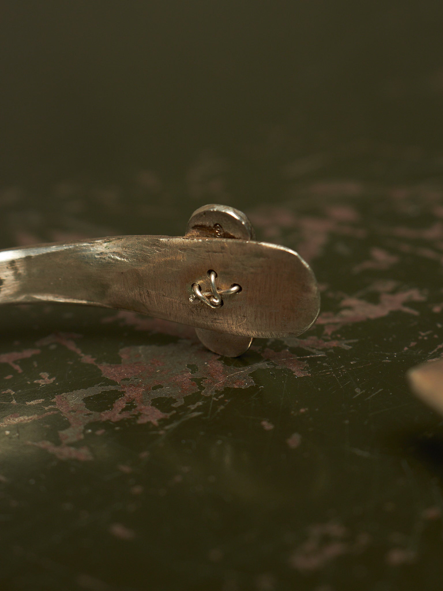 A close-up of a silver metal spoon handle with a small engraved anchor symbol, resting on a dark, textured surface.