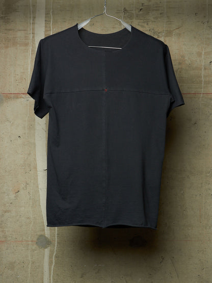 A dark charcoal gray short-sleeve t-shirt with a unique central seam and a tiny red dot detail, hanging on a metal hanger against a textured concrete wall.