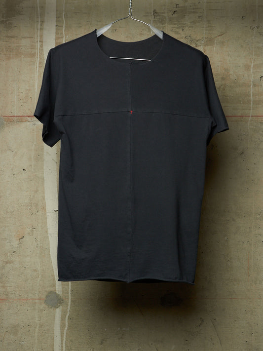 A dark charcoal gray short-sleeve t-shirt with a unique central seam and a tiny red dot detail, hanging on a metal hanger against a textured concrete wall.
