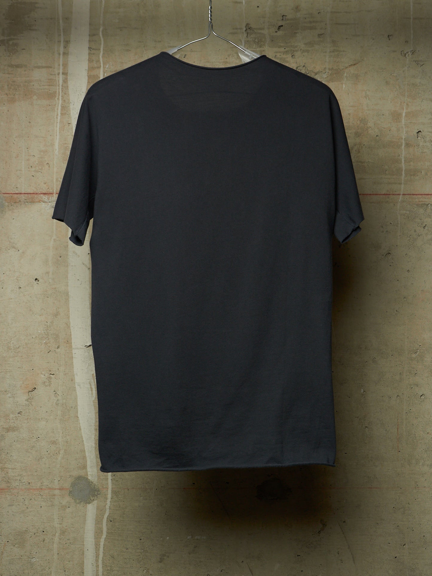 A dark charcoal gray short-sleeve t-shirt hanging on a clear plastic hanger against a textured concrete wall.