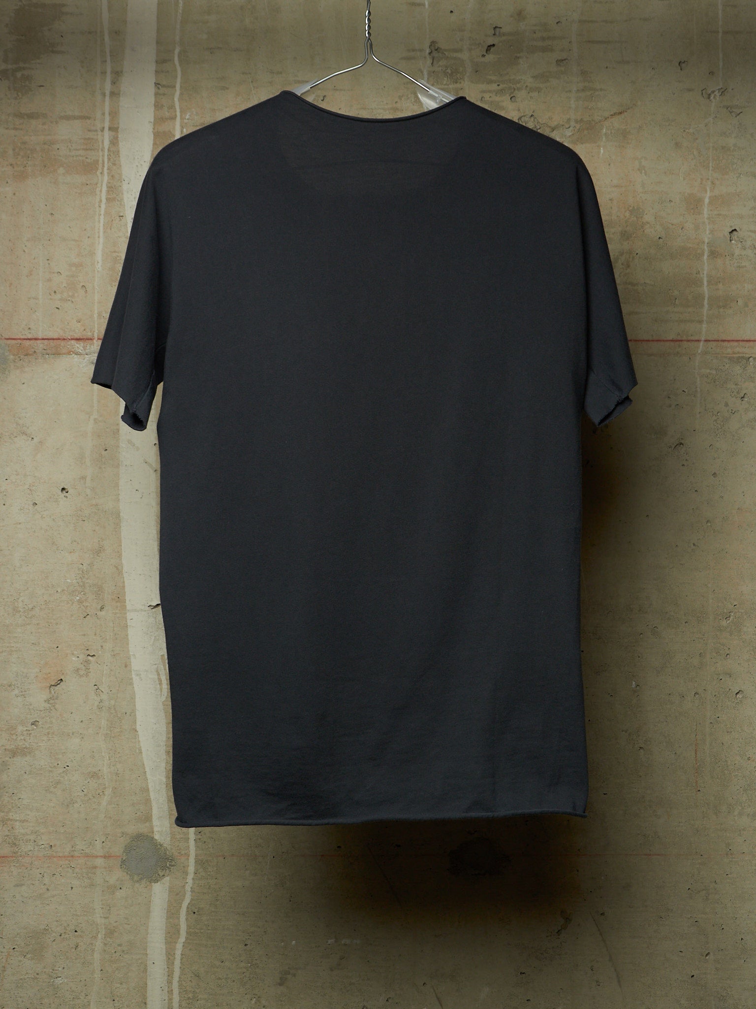 A dark charcoal gray short-sleeve t-shirt hanging on a clear plastic hanger against a textured concrete wall.