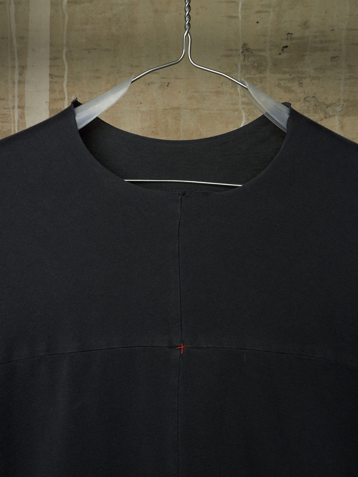 A black crewneck top hanging on a metal hanger against a textured concrete wall. The garment features a central vertical seam and a tiny red stitched cross detail on the chest.