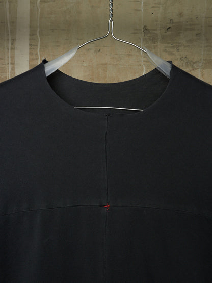 A black crewneck top hanging on a metal hanger against a textured concrete wall. The garment features a central vertical seam and a tiny red stitched cross detail on the chest.