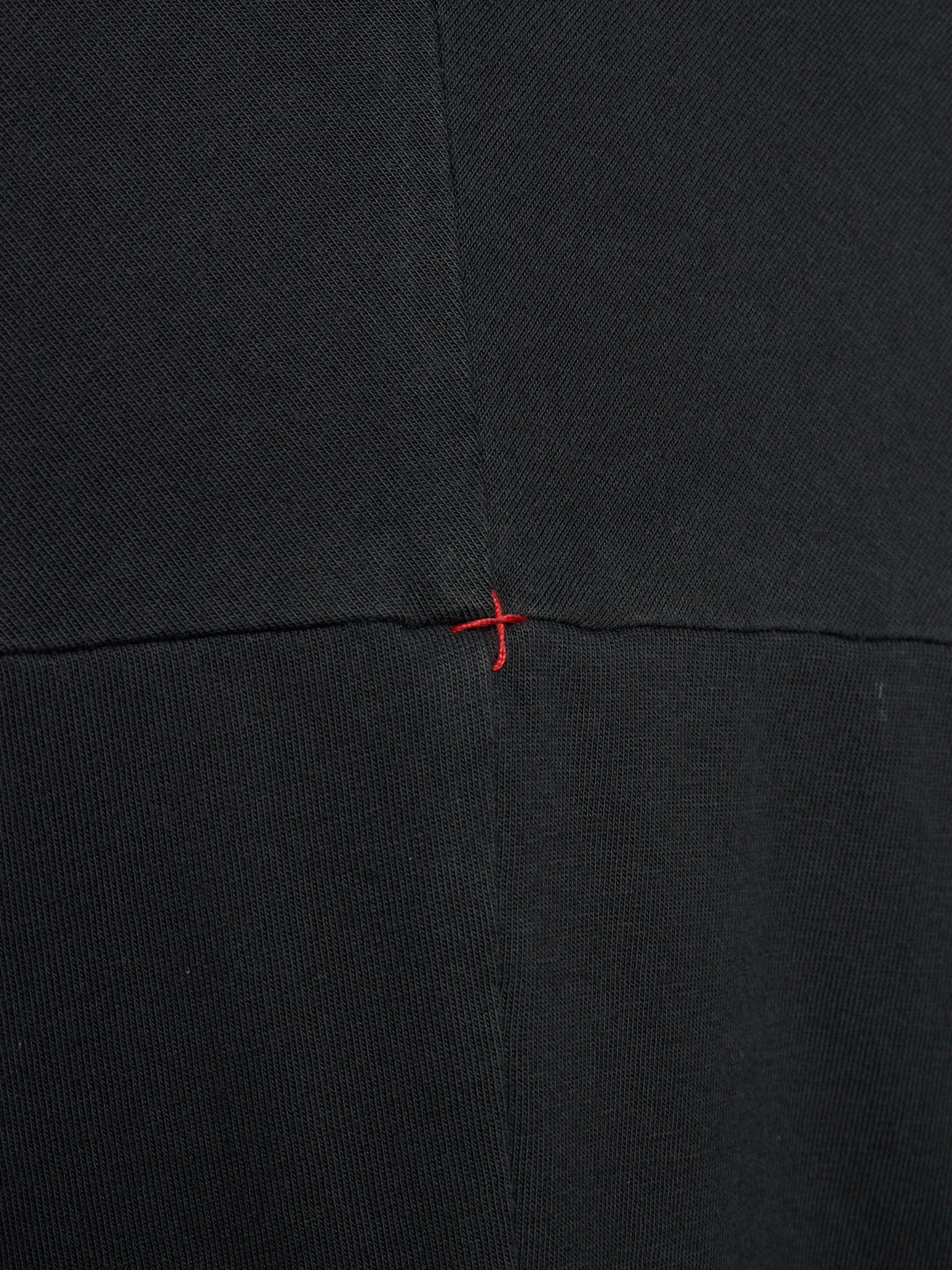 Close-up of a black textured fabric with a central horizontal seam and a small red stitched cross detail at the intersection.