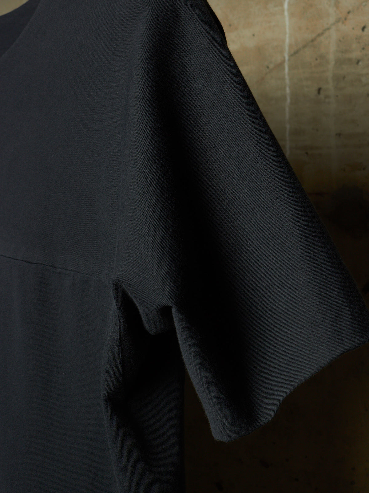 Close-up of a black short-sleeve top with a relaxed, oversized fit, showing the fabric texture and a seam detail against a textured concrete background.