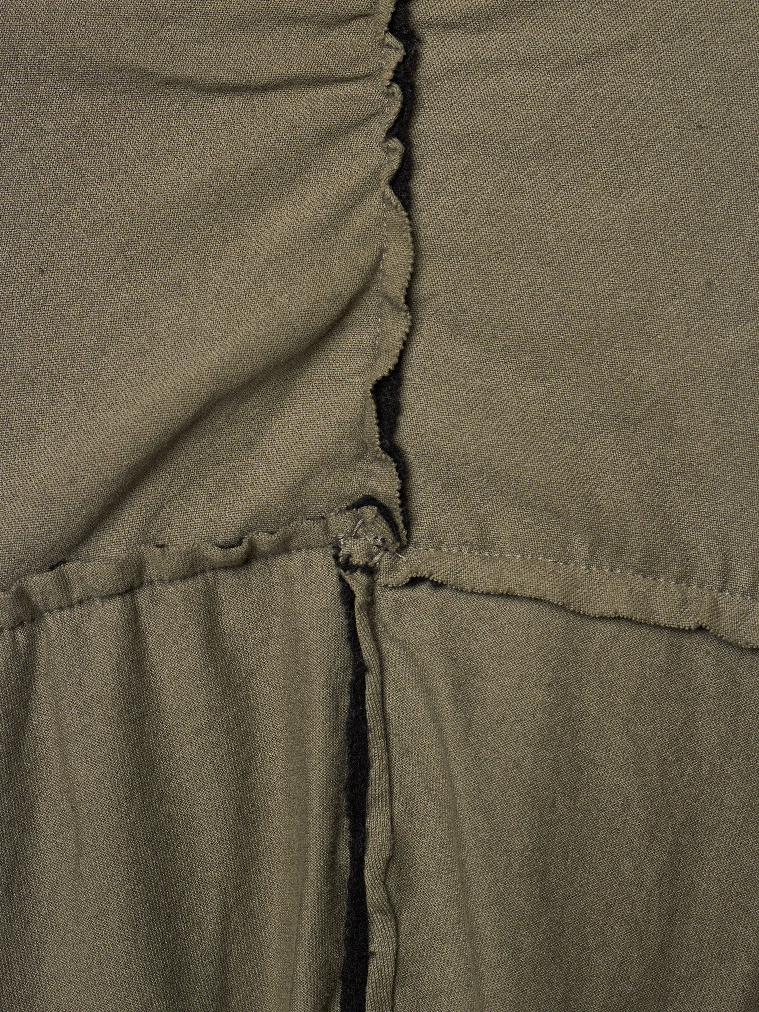 Close-up of olive green fabric with a gathered vertical seam and a horizontal stitched seam, showing the texture and construction details.