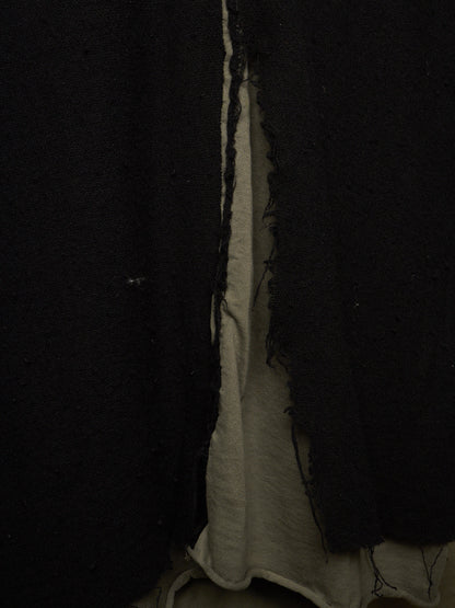 A close-up of a black textured garment, likely a cardigan or coat, featuring raw, frayed edges and a layered design with a light beige inner lining visible in the center.