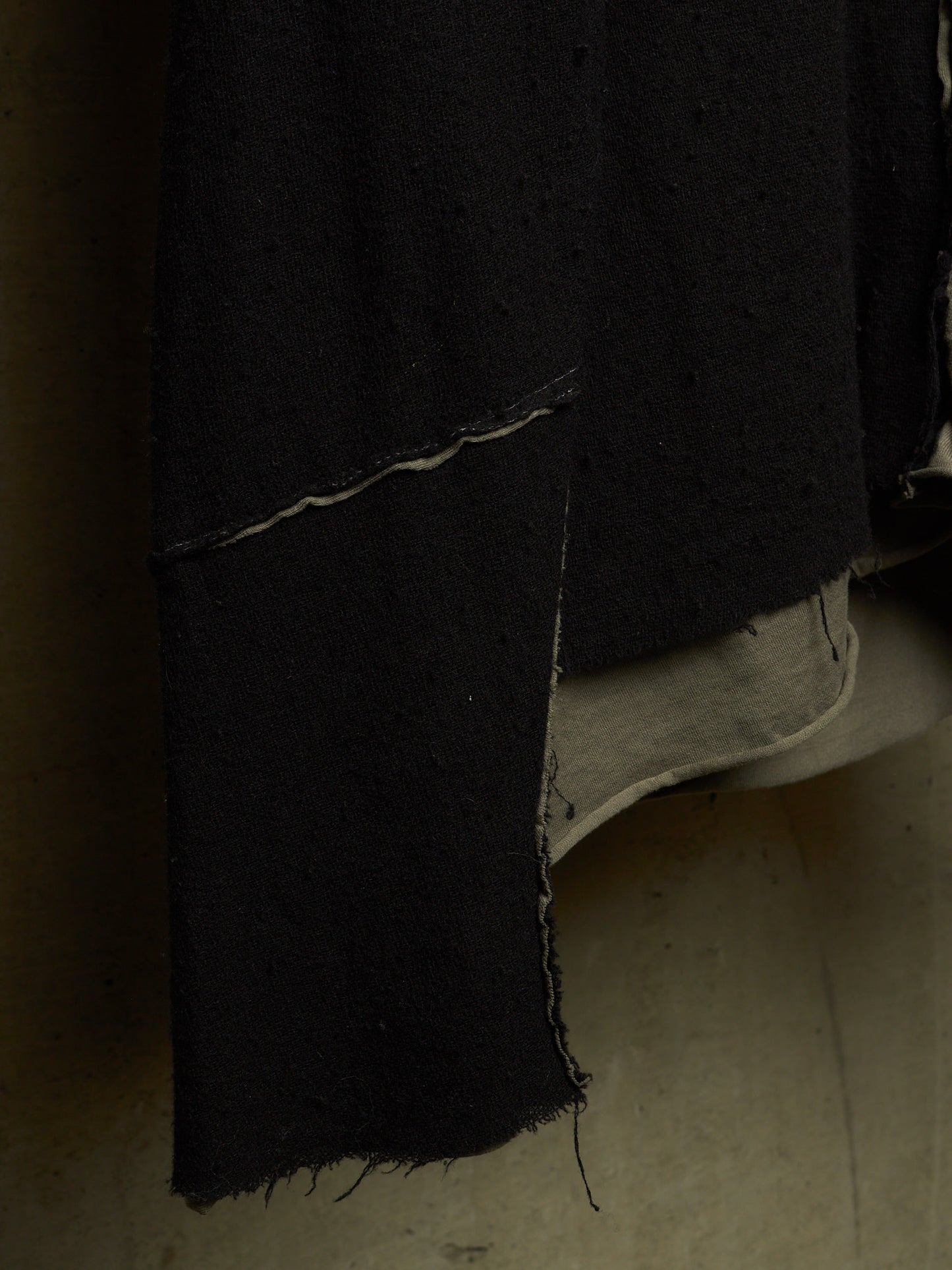 A close-up of a black textured garment or fabric panel with a raw, frayed hem, layered over a light beige underlayer, set against a dark, shadowed background.