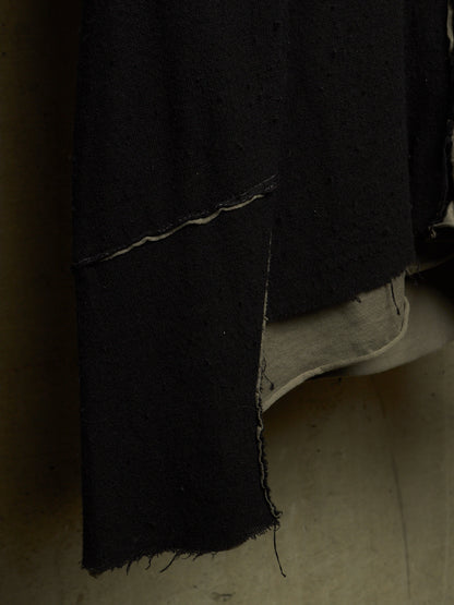 A close-up of a black textured garment or fabric panel with a raw, frayed hem, layered over a light beige underlayer, set against a dark, shadowed background.