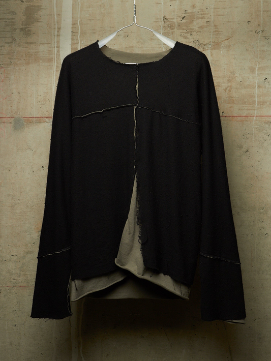 A black long-sleeve top with raw-edge seam detailing and a layered beige underlayer, hanging on a white wire hanger against a textured concrete wall.