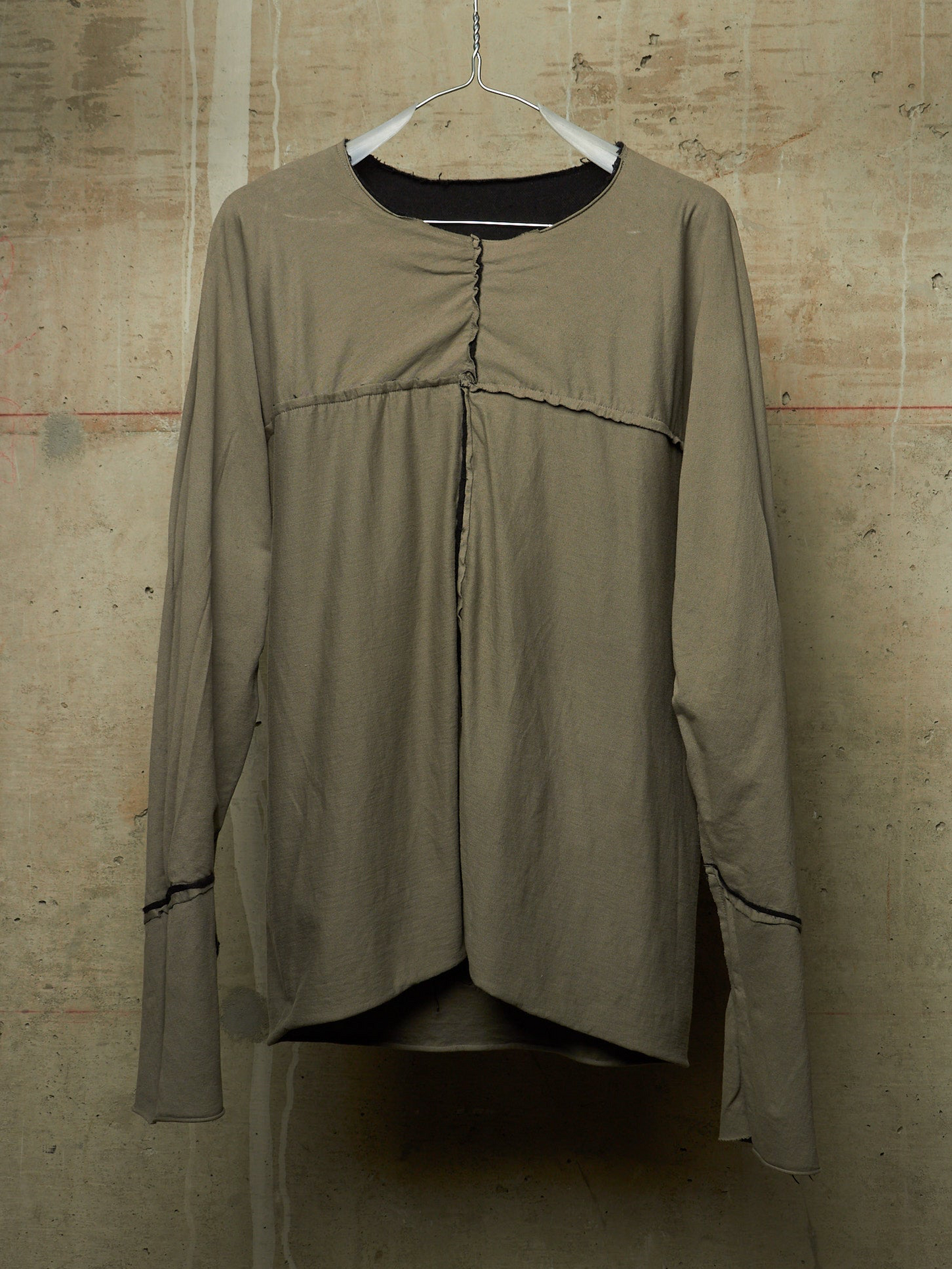 A long-sleeve top in a muted taupe or khaki color, hanging on a white hanger against a textured concrete wall. The top features a gathered front seam, a round neckline with a black inner collar, and thin dark contrast piping along the cuffs.