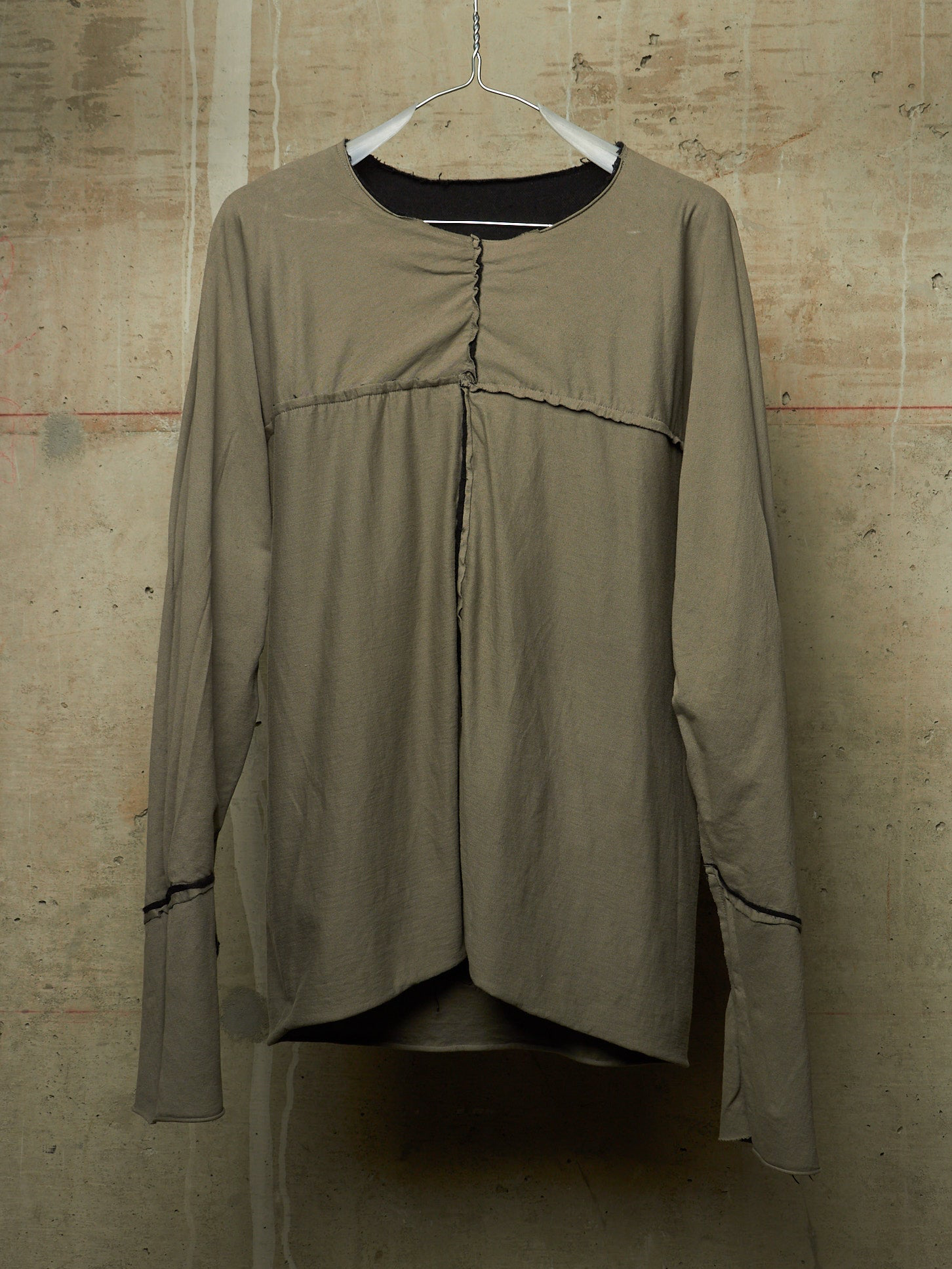 A long-sleeve top in a muted taupe or khaki color, hanging on a white hanger against a textured concrete wall. The top features a gathered front seam, a round neckline with a black inner collar, and thin dark contrast piping along the cuffs.