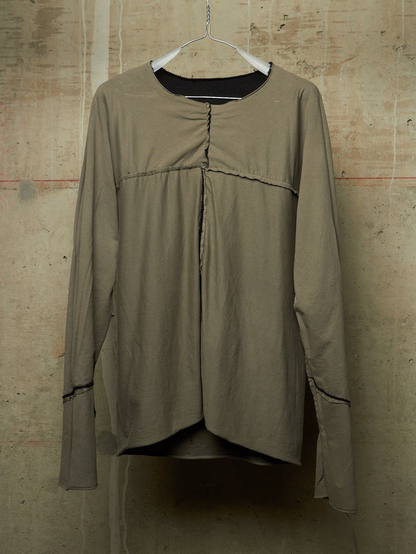 A long-sleeve top in a muted taupe or khaki color, hanging on a white hanger against a textured concrete wall. The top features a gathered front seam, a round neckline with a black inner collar, and thin dark contrast piping along the cuffs.