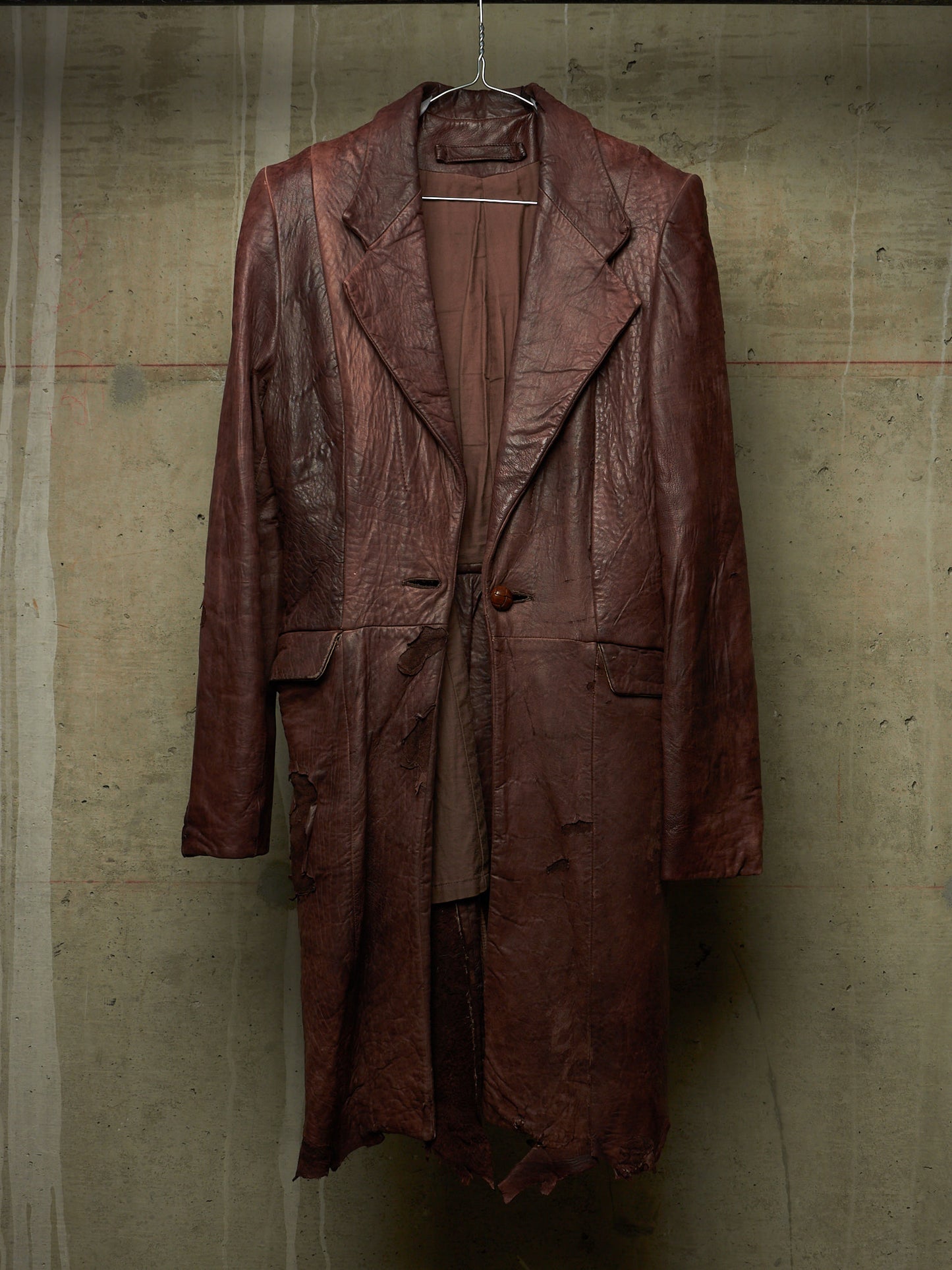 A distressed, heavily textured dark brown leather coat hanging on a wire hanger against a concrete wall.