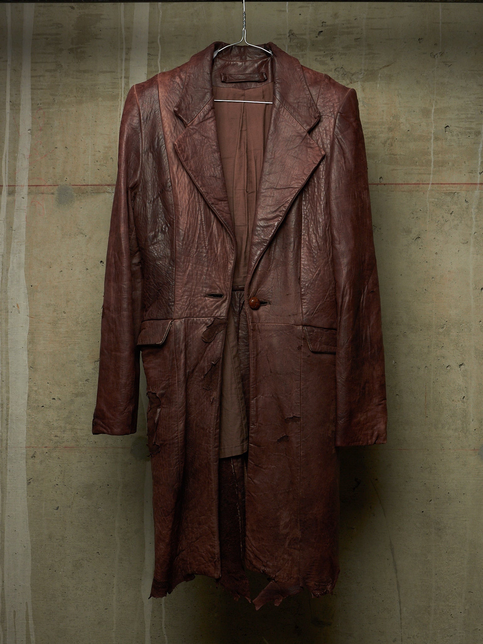 A distressed, heavily textured dark brown leather coat hanging on a wire hanger against a concrete wall.