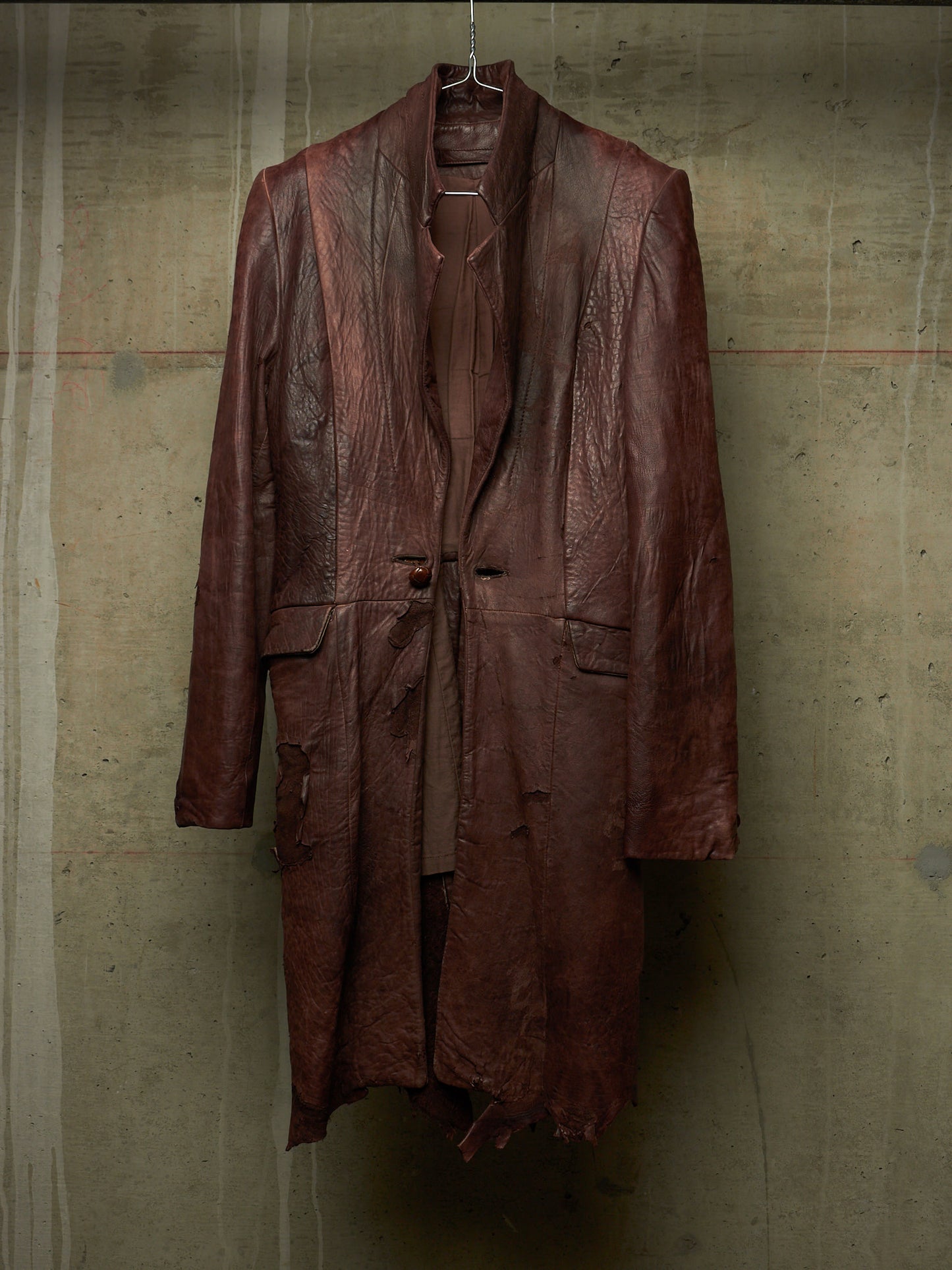 A distressed, dark reddish-brown leather coat hanging on a wire hanger against a textured concrete wall.