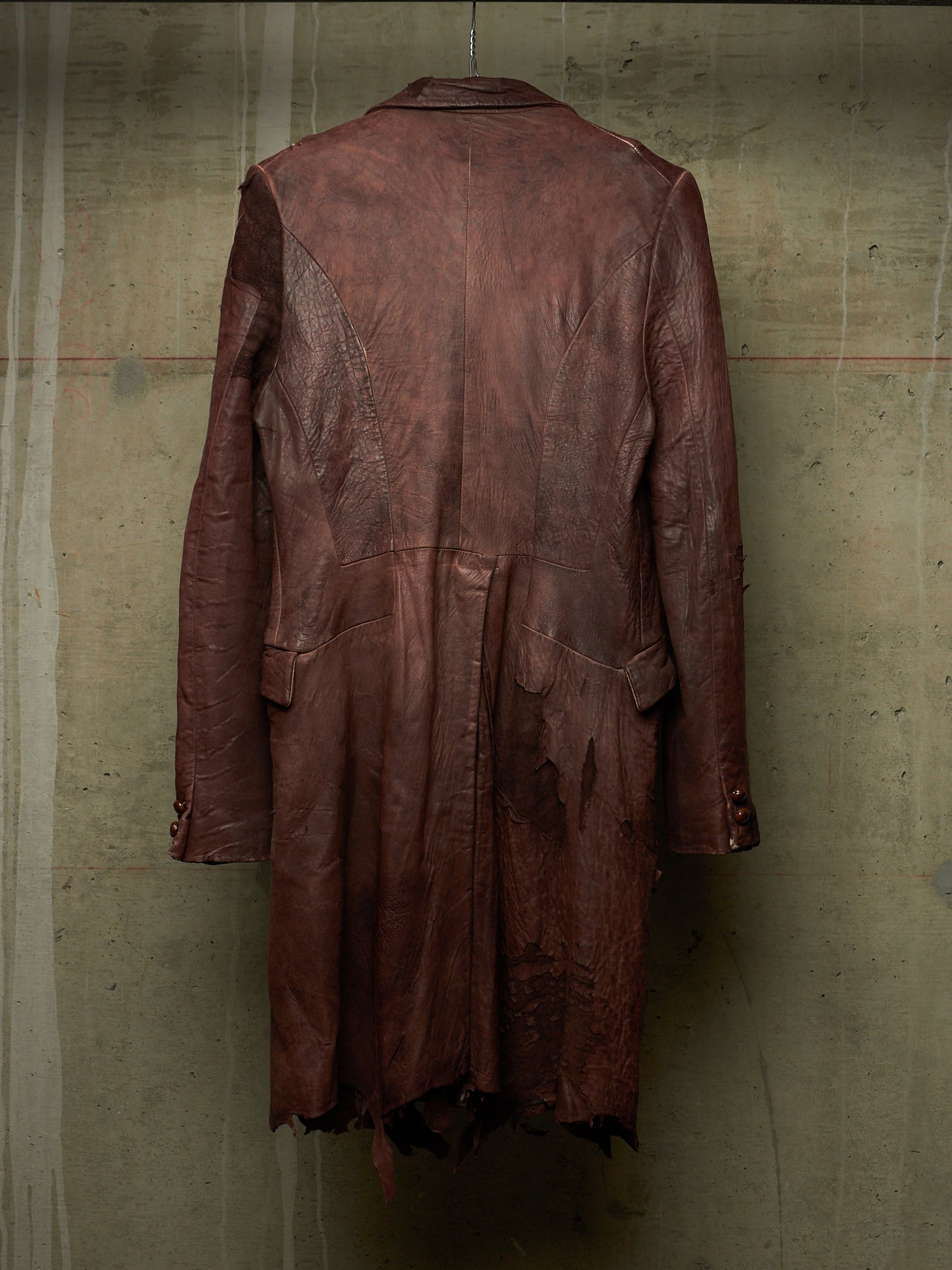 A distressed, dark reddish-brown leather coat hanging against a textured concrete wall, showing the back view with frayed, tattered details.