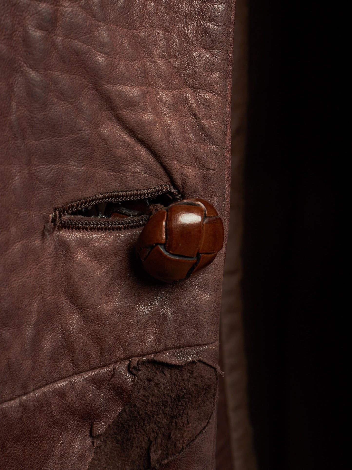 A close-up of a brown leather jacket featuring a unique, sculptural brown leather button on a zipper pocket, set against a dark background.