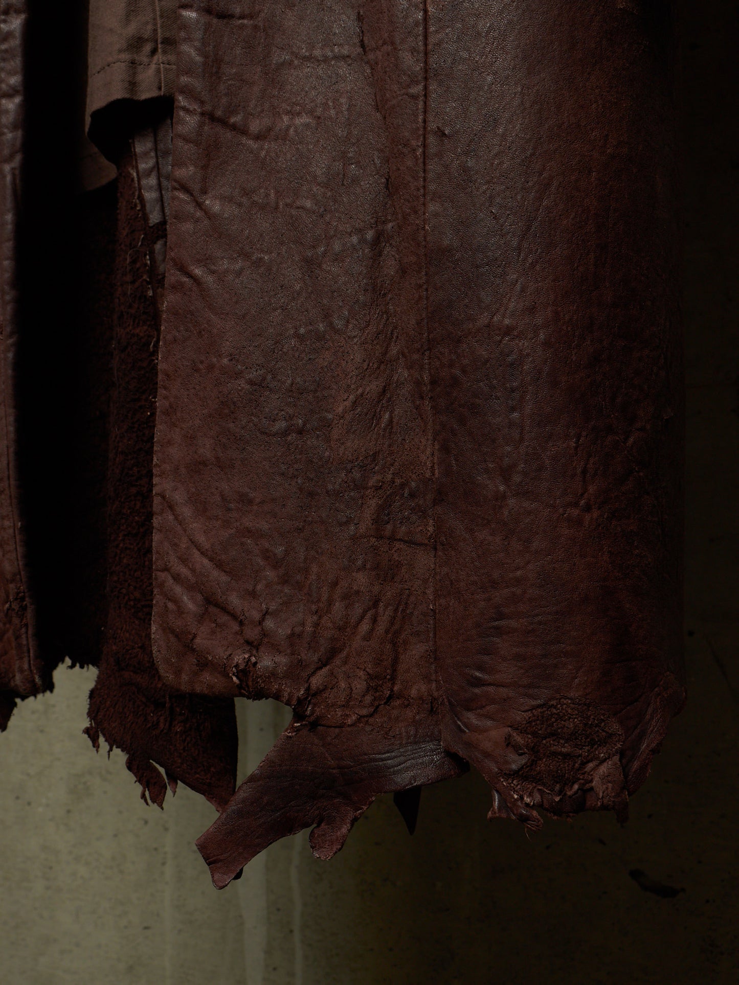 A close-up of dark brown textured leather or faux leather fabric hanging vertically, showing a wrinkled surface and frayed edges against a dim neutral background.