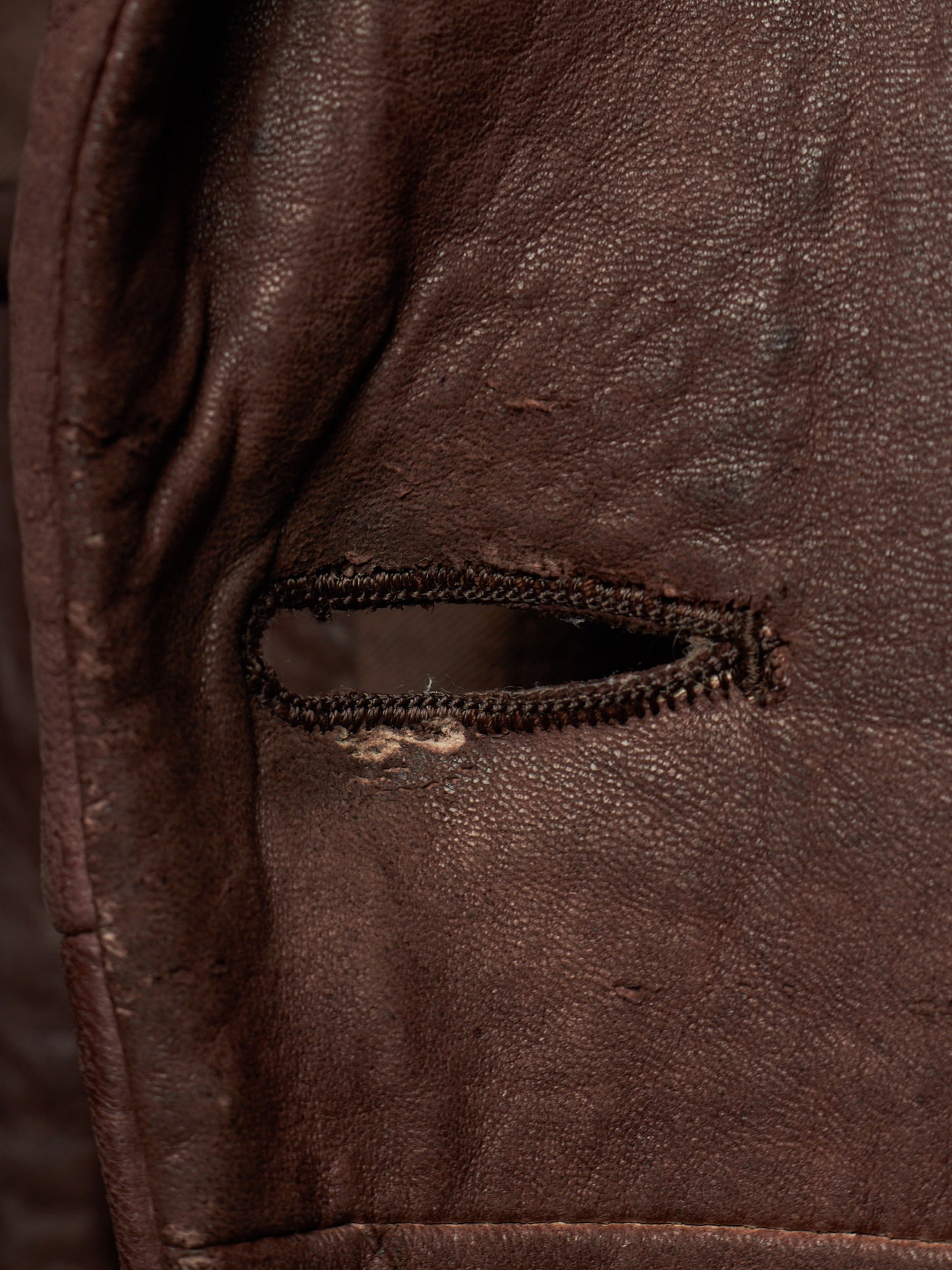 Close-up of a dark brown leather jacket showing a small zippered pocket with a worn zipper pull and visible fabric distressing.