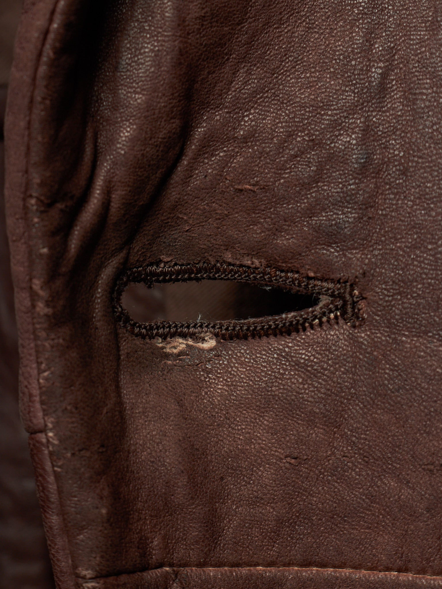 Close-up of a dark brown leather jacket showing a small zippered pocket with a worn zipper pull and visible fabric distressing.