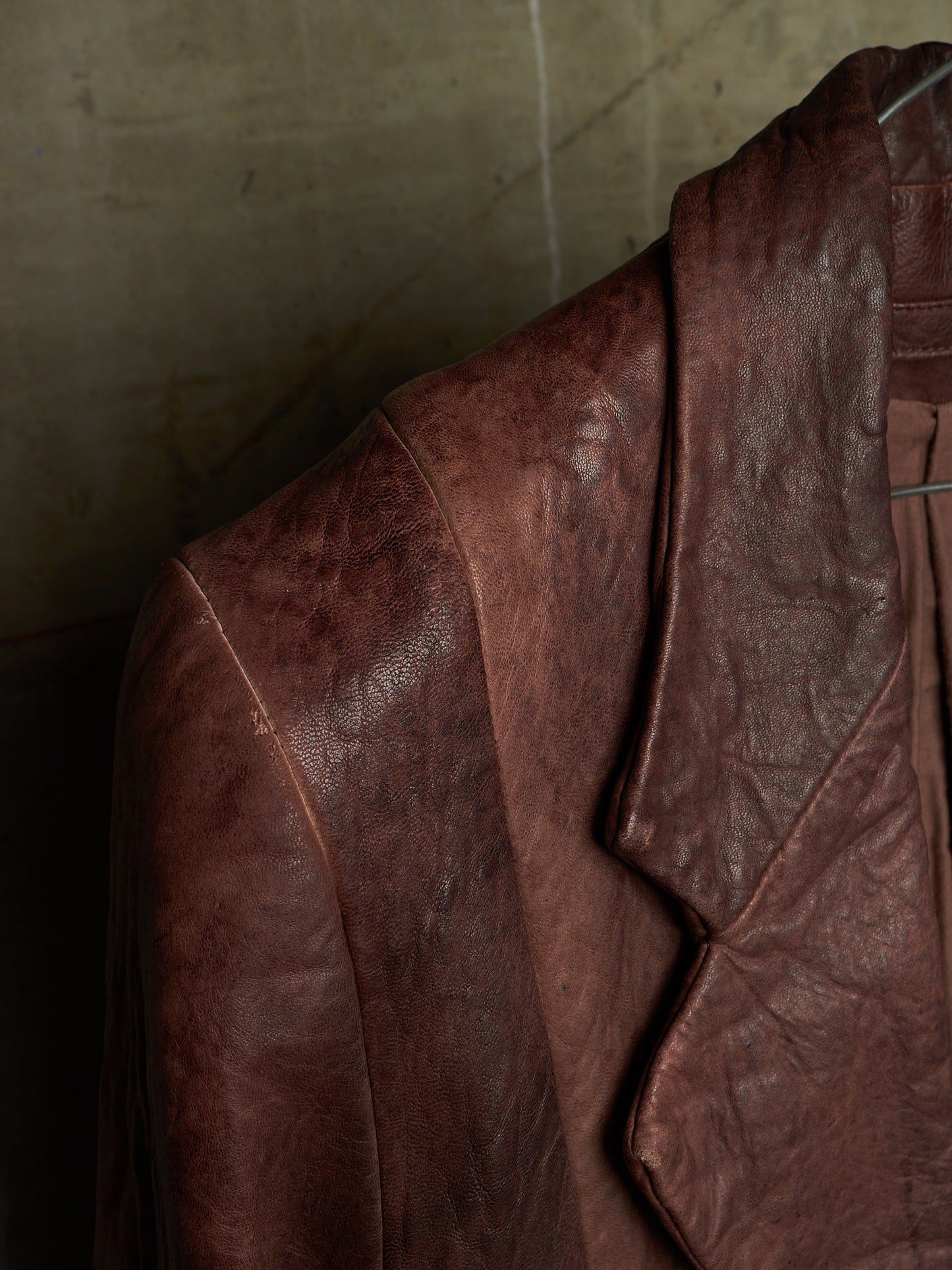 A close-up of a distressed brown leather jacket hanging against a textured concrete wall.