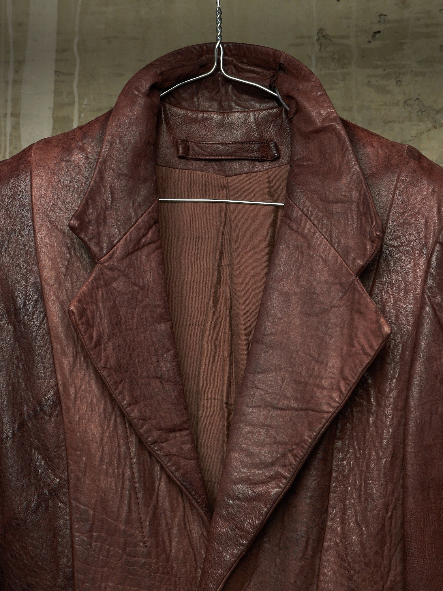 A close-up of a brown leather jacket hanging on a metal hanger against a textured concrete wall.