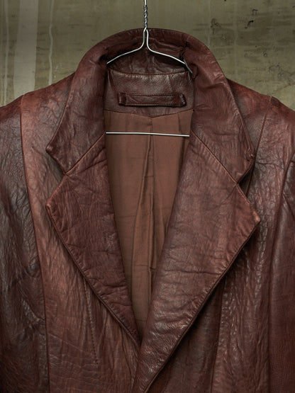 A close-up of a brown leather jacket hanging on a metal hanger against a textured concrete wall.