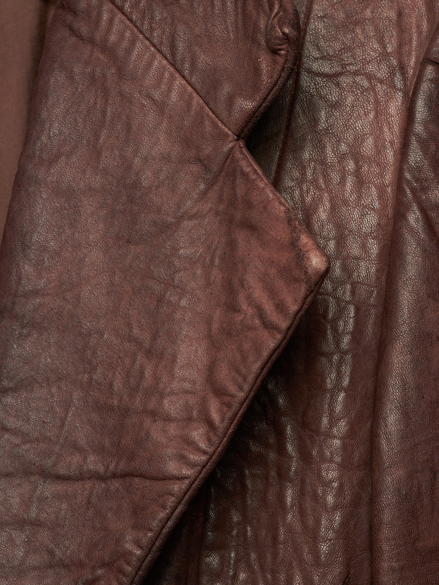 A close-up of a brown leather jacket showing the textured, crinkled leather material and a diagonal fold.