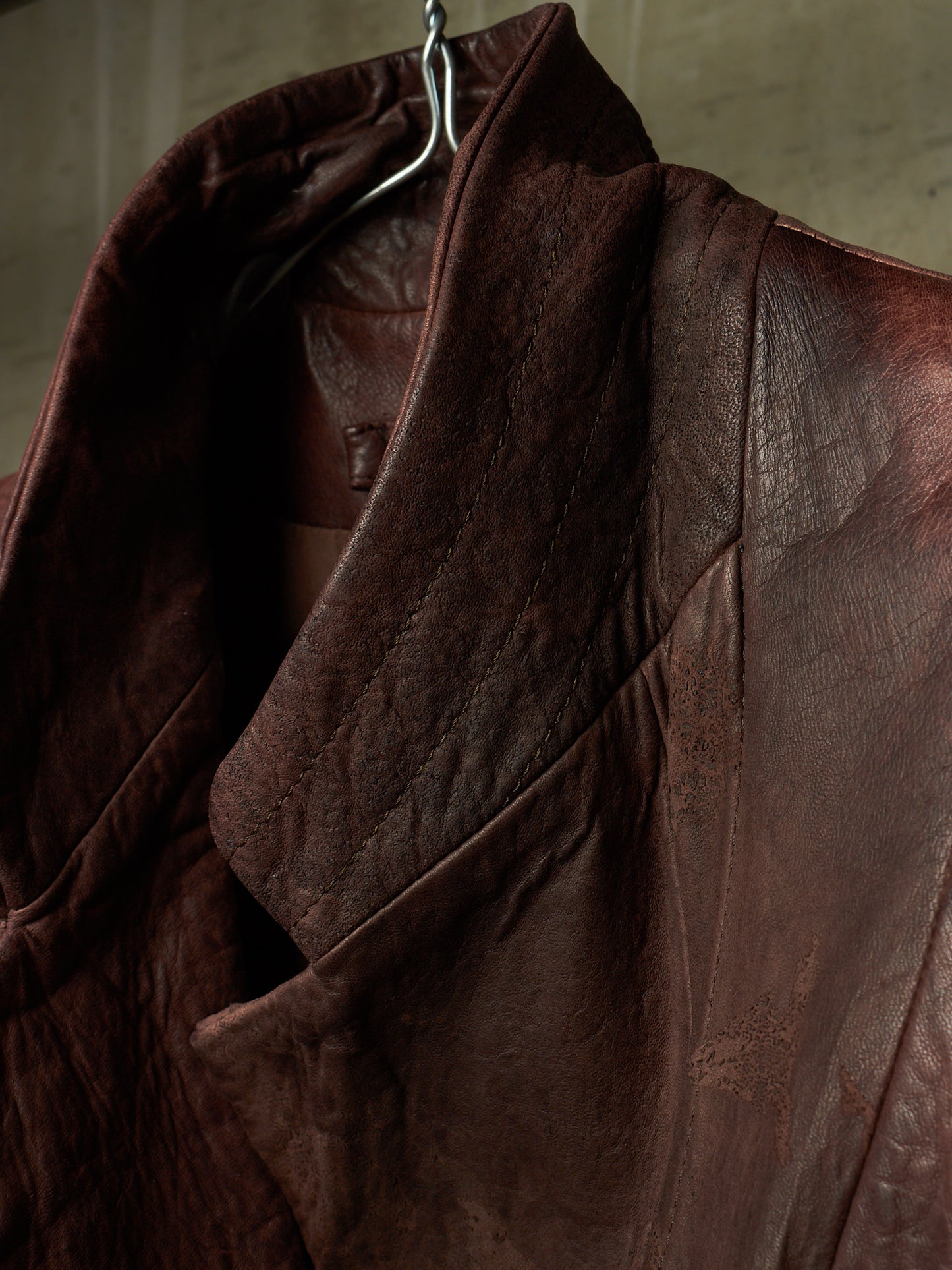 A close-up of a dark brown leather jacket hanging on a metal hanger against a textured gray wall.