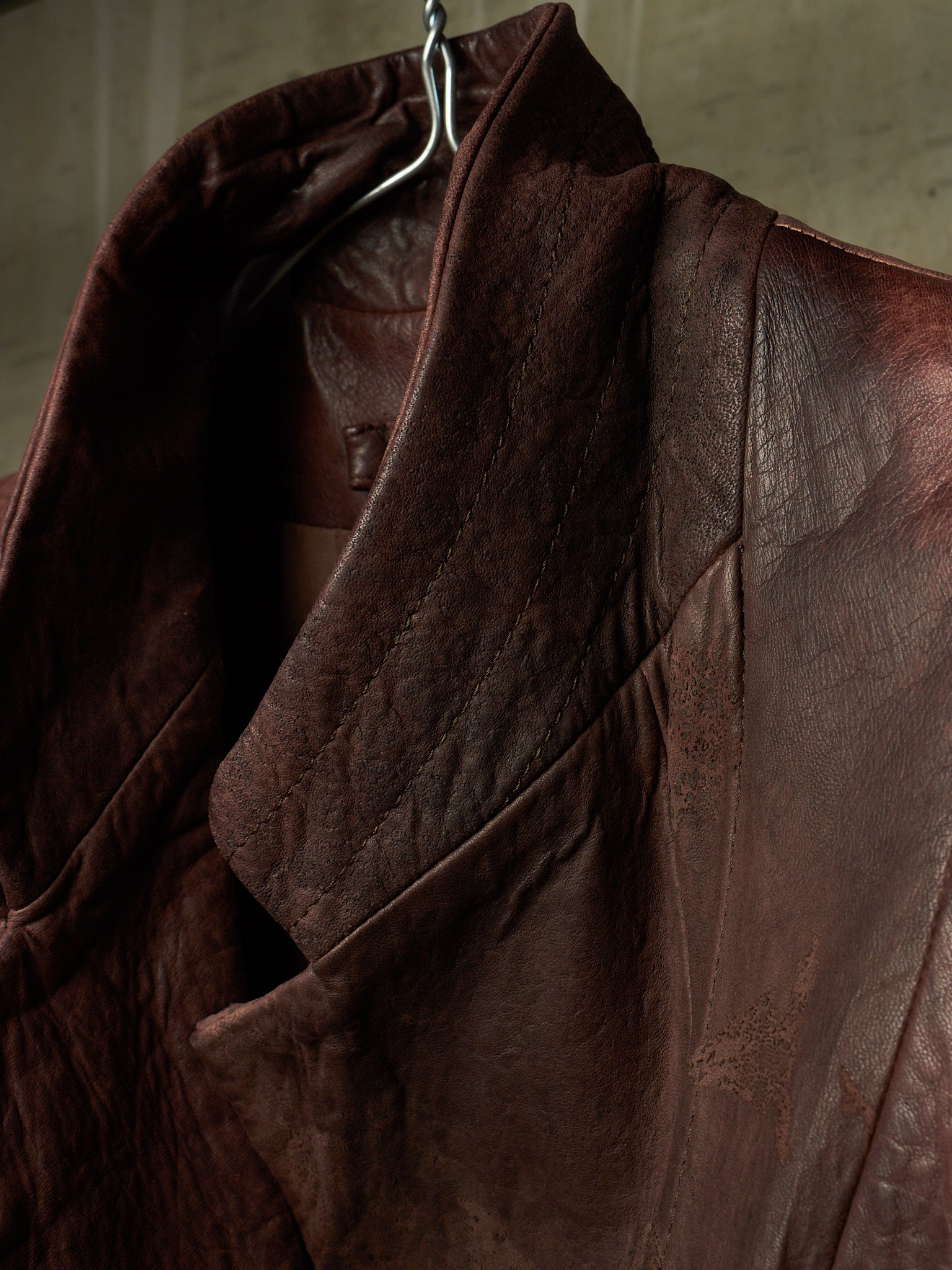 A close-up of a dark brown leather jacket hanging on a metal hanger against a textured gray wall.