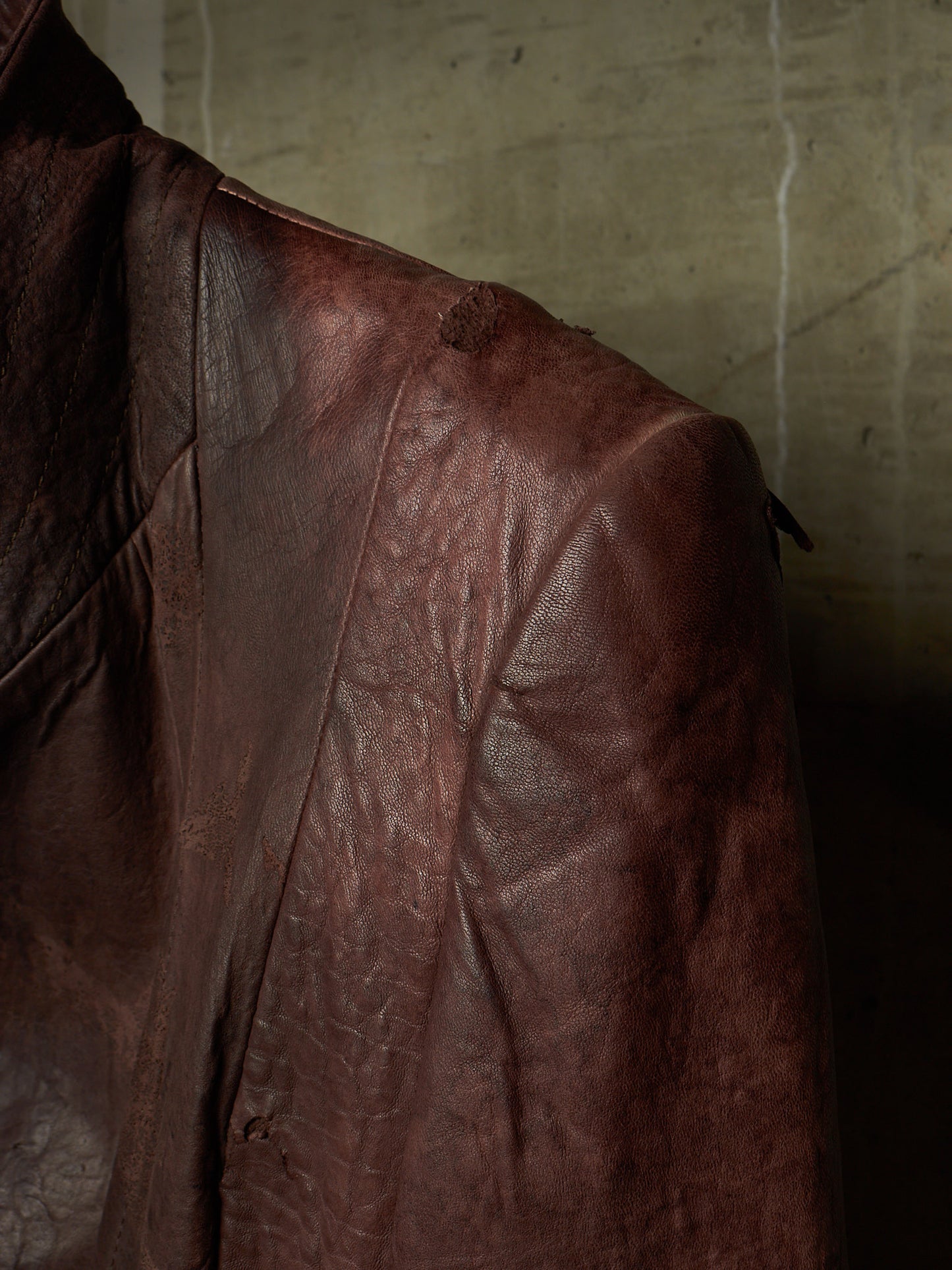 A close-up of a distressed brown leather jacket, showing the textured leather fabric and shoulder detail against a dark, moody background.