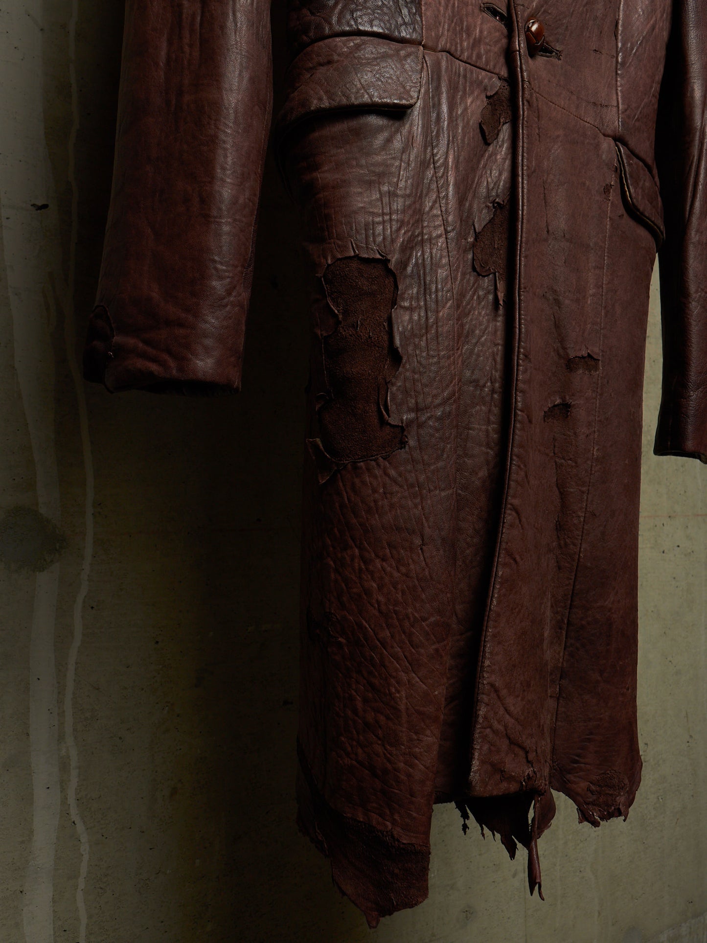 A dark brown distressed leather coat with heavy rips and tattered edges, hanging against a textured concrete wall.