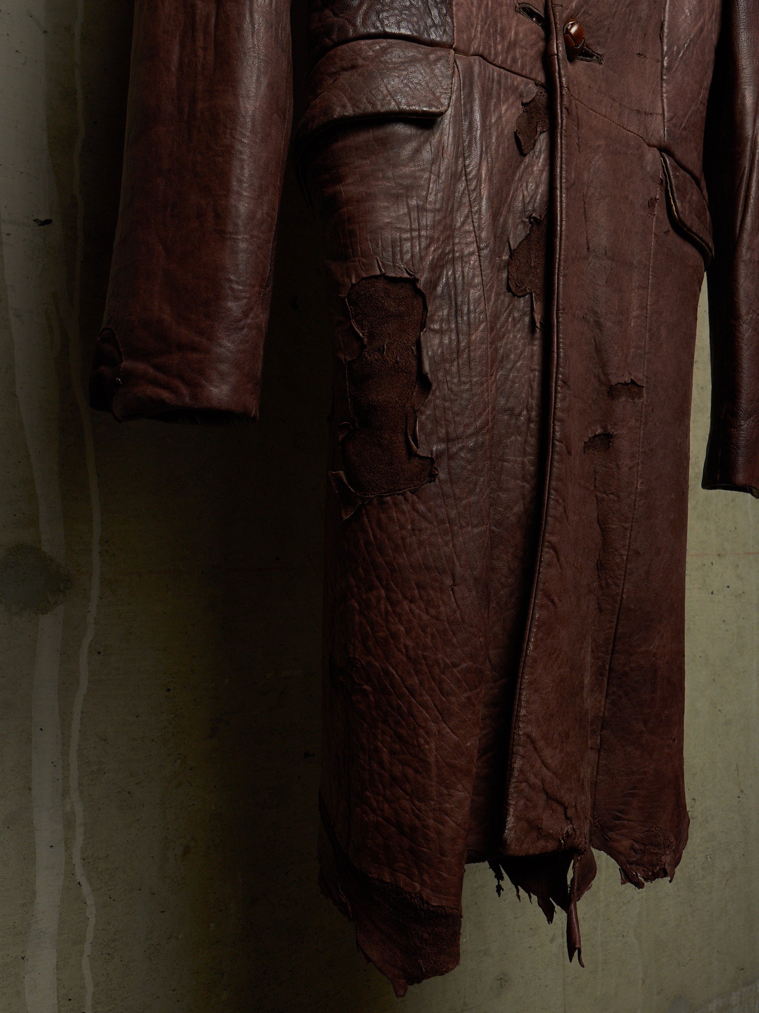 A dark brown distressed leather coat with heavy rips and tattered edges, hanging against a textured concrete wall.