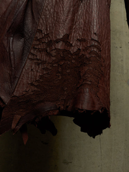 A close-up of a dark reddish-brown leather garment or material with heavily distressed, cracked, and shredded texture along the lower edge, set against a plain concrete-like gray background.
