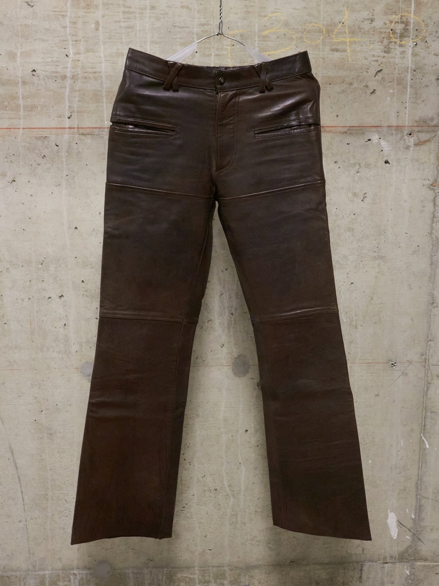 Brown leather pants on a hanger against a textured wall.