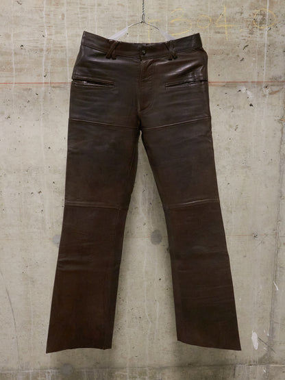 Brown leather pants on a hanger against a textured wall.