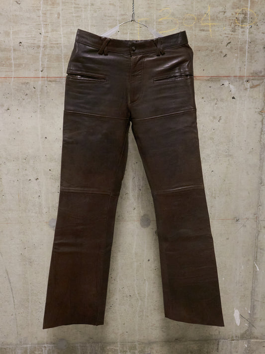 Brown leather pants on a hanger against a textured wall.