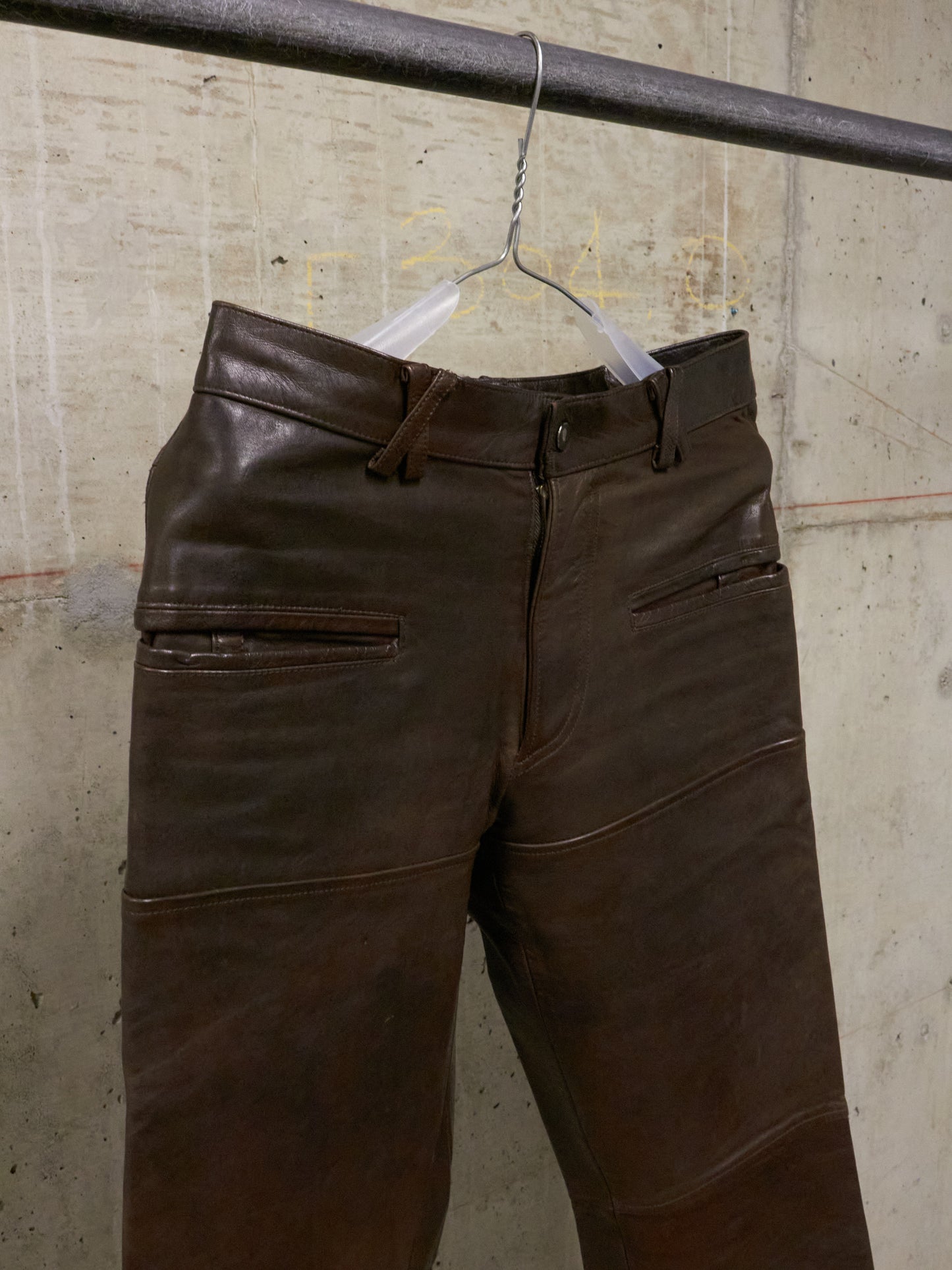 Brown leather pants hanging on a hanger against a textured wall.