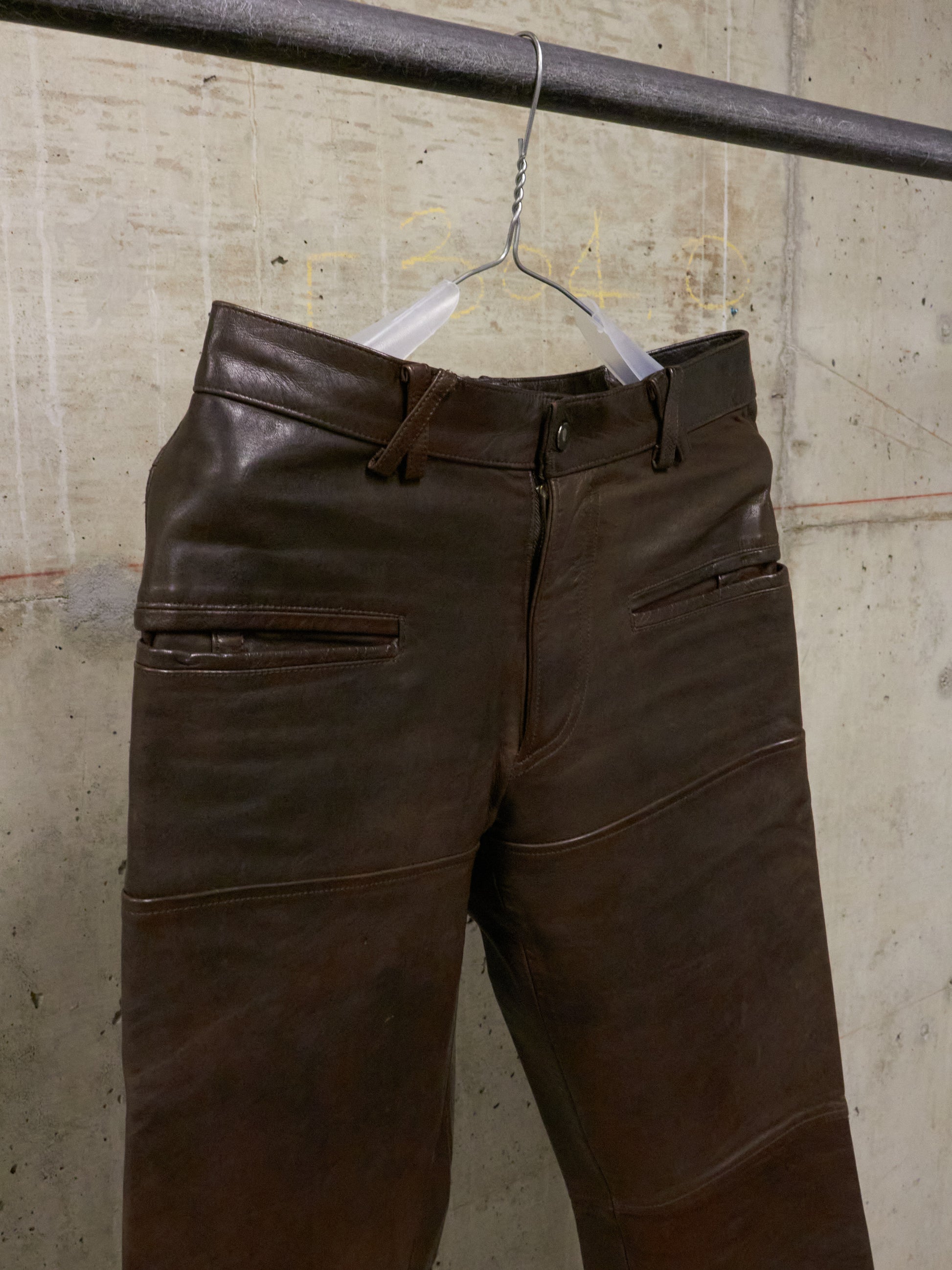 Brown leather pants hanging on a hanger against a textured wall.