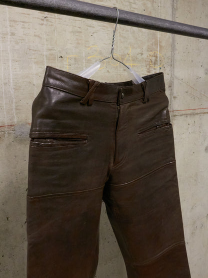 Brown leather pants hanging on a hanger against a textured wall.